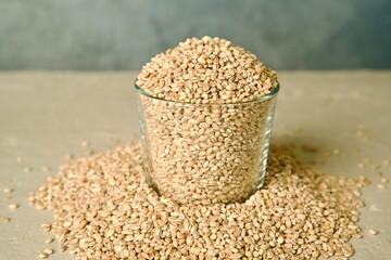 A glass jar filled with pearl barley, with scattered barley grains around it on a light surface.