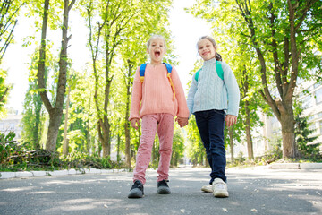 Obraz premium Two girls, full of laughter and energy, stroll hand in hand along a sunlit road framed by lush green trees, enjoying their time after school.