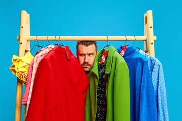 Curious man squeezed between bright clothes on rack, peering cautiously while eyeing discounts...