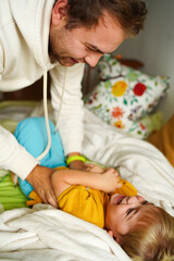 A father playfully tickles his laughing child on the bed, surrounded by blankets, capturing a...