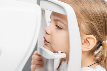 A pediatric optometrist examines the eyes of a little girl using modern equipment. The happy girl...