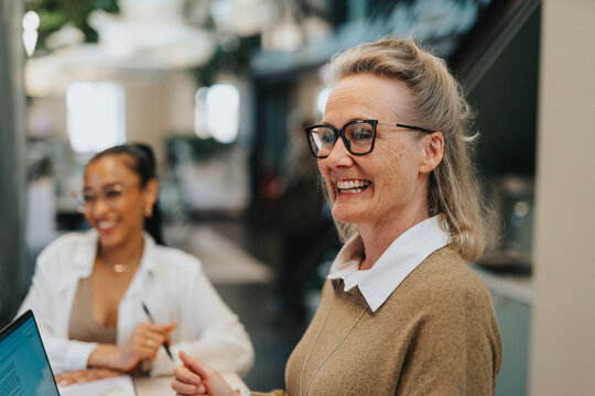 Happy businesswoman wearing eyeglasses working on laptop with coworker in office