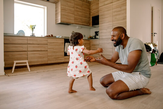 Baby girl taking first steps near father sitting on floor at home