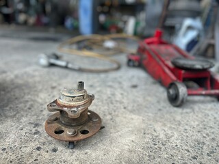 A close up of a rusty automotive part on a garage floor, evoking a sense of nostalgia and mechanical work. The background features tools and a jack, indicating an active workspace.