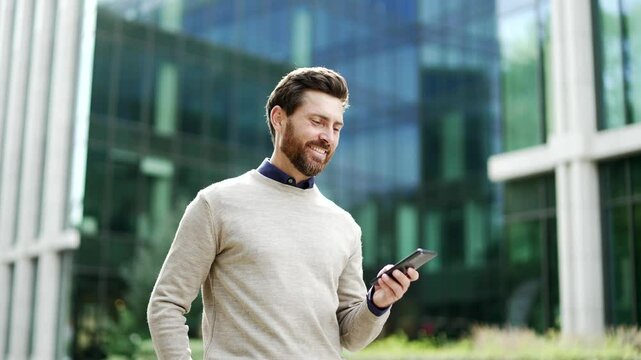 Businessman walking outdoors holding smartphone, smiling while browsing or chatting online. Male professional in casual enjoying city life, checking messages on street near business office building