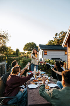 Happy young woman doing celebratory toast with male and female friends sitting near dining table for dinner party in bac