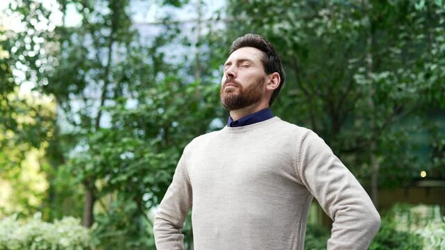 Mature male professional standing in city park, eyes closed, taking deep breath. Concept of relaxation, mindfulness, and stress relief in nature. Businessman appreciating serene outdoor environment