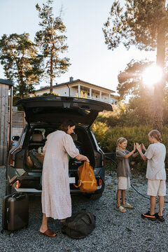 Mother loading luggage with boys playing clapping game while standing near car trunk on driveway