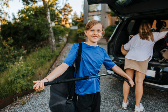 Portrait of smiling boy holding ice hockey stick with mother loading car trunk