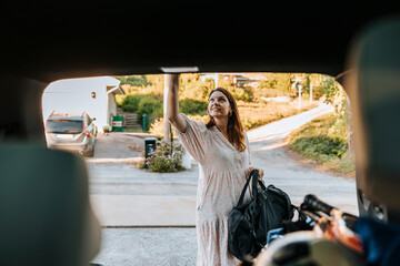 Mature woman holding bag and closing car trunk while standing on driveway