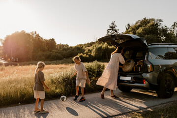 Mother near car trunk with boys playing with soccer ball on driveway by meadow at sunny day