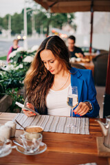 Young beautiful adult woman wearing jacket sitting next to the table in street cafeteria and hold smart phone device and glass of sparkling wine drink romantic date or businesswoman rest