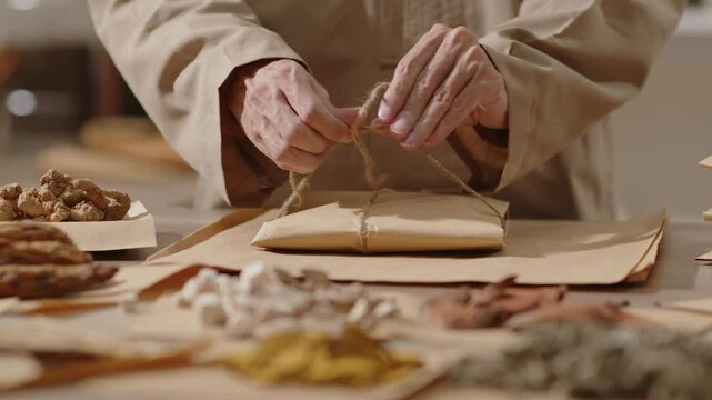 A herbalist is tying a package of herbs with a beige string, while other herbs are placed in wrapping paper. Video template with a traditional medicine room background for content traditional medicine