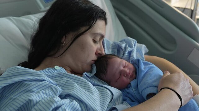 Mother kissing newborn baby infant right after birth, initial day of infant being held and cared by mom at hospital clinic cradling son in arms