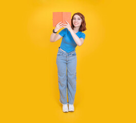 Full body size caucasian red bob hair woman wear blue t shirt and jean over yellow studio background playfully hides behind an open book toothy smile looking camera, copy space, mock up.