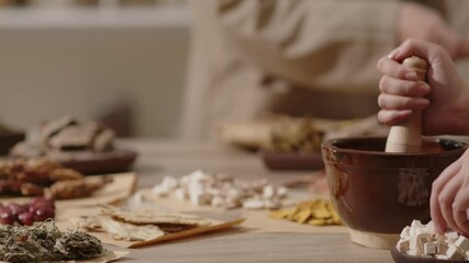 The Oriental Medicine room setting with a table filled with herbs features a herbalist pounding angelica sinensis while a herbalist in beige clothes rearranges the wrapped herbal medicine packets.