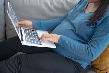 Fototapeta premium Pregnant asian woman using laptop sitting on sofa at home buying for newborn Online shopping pregnancy concept Positive life moment in pregnancy time.