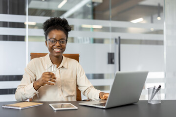 Businesswoman sits at office desk holding glass water, smiling while working on laptop. Smartphone...