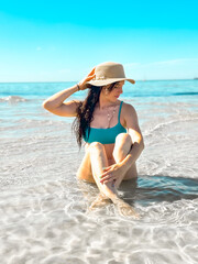 A young  woman in a swimsuit and hat sits in the ocean waves on a sunny Melbourne beach, Sorrento. Enjoying a peaceful summer day, she relaxes by the coast, embracing the beauty of nature and freedom