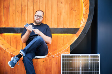 Bearded man connecting smartphone to photovoltaic solar panel. Integration of sustainable renewable...