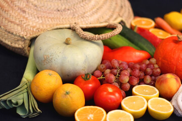 Round straw bag and various seasonal fruits and vegetables on dark background. Summer and fall produce. Selective focus.