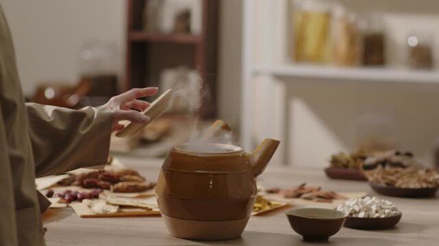 A woman reaches for a wooden lid to reveal a pot filled with steaming herbal tea, a traditional Chinese medicine drink, on a wooden table