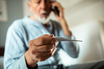 Elderly man checking thermometer while calling doctor at home