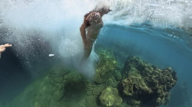 A woman captured mid-jump as she dives from a cliff into the water, with bubbles forming around her as she descends and rocks visible below.