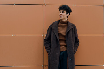 Fashionable young man in a brown coat and turtleneck sweater, smiling with hands in pockets, leaning against a wall