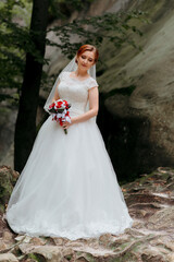 Fototapeta premium A woman in a white dress is holding a bouquet of red flowers. She is standing on a rocky surface, possibly a mountain or a cliff. Concept of natural beauty and serenity