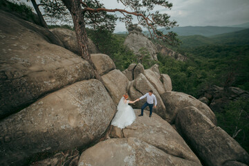 A bride and groom are climbing a rocky mountain together. The bride is wearing a white dress and the groom is wearing a blue shirt. The scene is peaceful and romantic