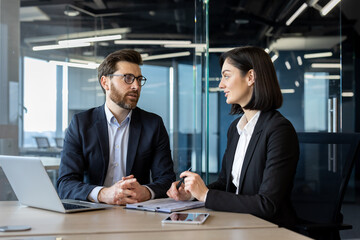 Two business professionals engaged in discussion across table with laptop and documents in...