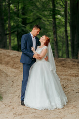 A bride and groom are standing in a forest, with the bride wearing a white dress and the groom wearing a blue suit