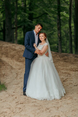 A bride and groom are posing for a picture in a forest. The bride is wearing a white dress and the groom is wearing a blue suit. Scene is happy and romantic