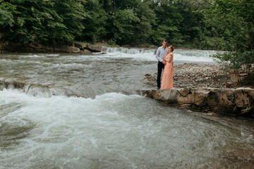 A couple standing on a rock near a river. The man is wearing a tie and the woman is wearing a pink dress