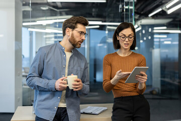 Two office workers engaged in conversation, reviewing project details on tablet. Man with coffee cup intently listening to woman explaining concepts. Modern workplace with glass walls