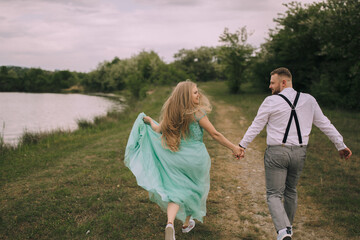 A couple is running together in a grassy field near a lake. The woman is wearing a green dress and the man is wearing a white shirt and black suspenders. Scene is joyful and romantic