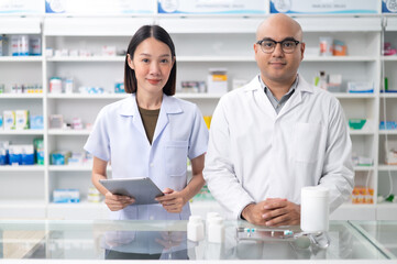Fototapeta premium Asian professional male pharmacist using tablet checking drug list holding pill bottle A pharmacist checks the stock of medicines on the shelf with a female pharmacist standing and giving advice.