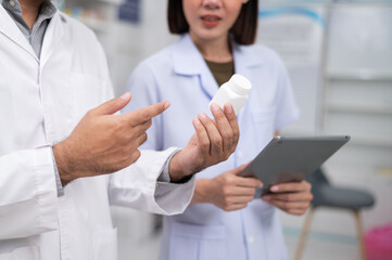 Fototapeta premium Asian professional male pharmacist using tablet checking drug list holding pill bottle A pharmacist checks the stock of medicines on the shelf with a female pharmacist standing and giving advice.