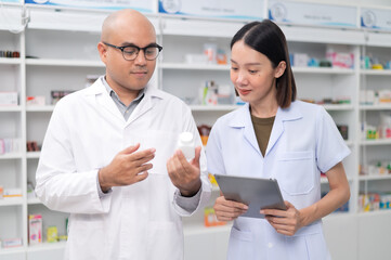 Obraz premium Asian professional male pharmacist using tablet checking drug list holding pill bottle A pharmacist checks the stock of medicines on the shelf with a female pharmacist standing and giving advice.