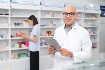 Fototapeta premium Asian professional male pharmacist using digital tablet computer checking drug list Pharmacist checking stock in pharmacy for health medicine in pharmacy with female pharmacist in the background
