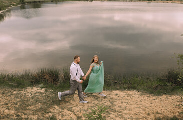 A couple is walking by a lake, with the man holding the woman's hand. The scene is peaceful and romantic, with the couple enjoying each other's company in a beautiful natural setting