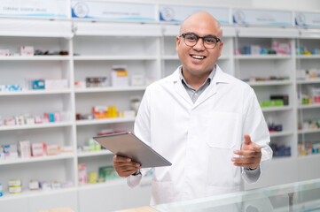 Asian professional male pharmacist using tablet checking drug list holding pill bottle Pharmacist checking stock in pharmacy on shelf looking at camera charming smile
