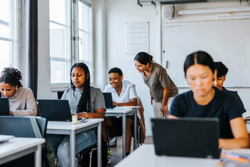 Smiling female teacher assisting student studying on laptop while sitting in classroom at school