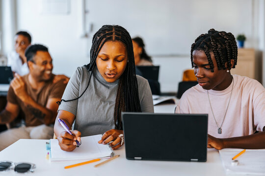 Braided hair girl writing while sitting beside male classmate near desk in classroom at school
