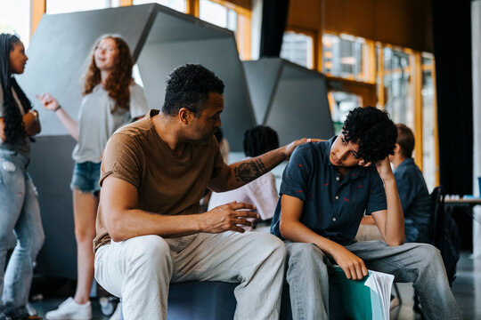 Male teacher consoling teenage boy leaning on elbow while sitting in cafeteria at school
