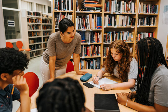 Female librarian assisting teenage students sitting near table in library at school