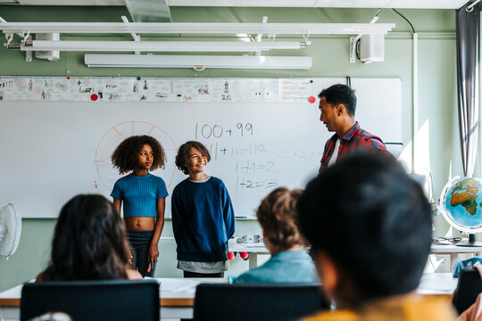 Teacher talking with male and female students standing near whiteboard in classroom
