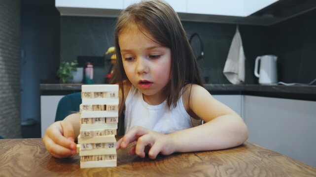 Table game, A cheerful child girl 5s happily plays all by herself with a wooden block tower game in cozy kitchen, showcasing her imaginative spirit as she builds her dreams, aspirations through play
