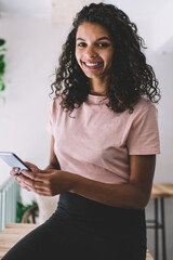 Half length portrait of dark skinned young woman looking at camera while sitting with cellphone indoors, African American smiling hipster girl holding modern mobile phone in hands during rest